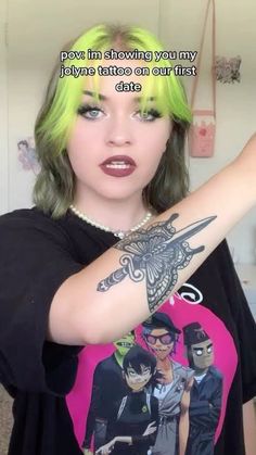 a woman with green hair and tattoos on her arm pointing at the camera while wearing a black t - shirt