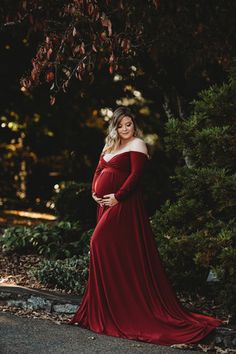 a pregnant woman in a red gown poses for a photo while leaning against a tree