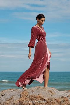 a woman standing on top of a rock next to the ocean wearing a pink dress