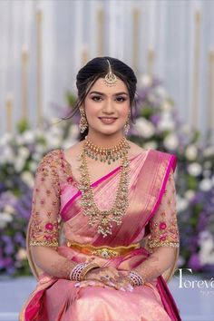 a woman in a pink and gold sari sitting on a blue surface with flowers behind her