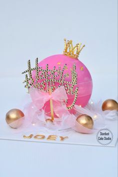 a pink cake with a tiara on top and some gold eggs in front of it