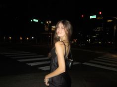 a woman walking across a street at night with her hand on her hip and wearing a black dress