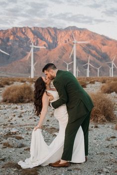 a bride and groom kissing in front of windmills at sunset with mountains in the background