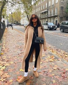 a woman is standing on the sidewalk with her hand in her pocket and wearing a tan coat