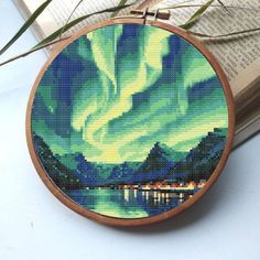 a cross - stitch aurora bore in the night sky with green and yellow lights, on top of an open book