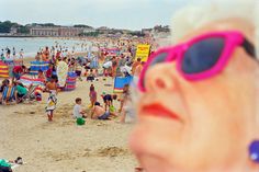 a woman wearing sunglasses on the beach with people in the water and sand behind her