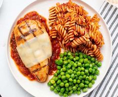 a white plate topped with pasta and meat covered in sauce next to green peas on a striped table cloth