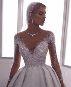 a woman in a wedding dress with pearls on the neck and long sleeves, standing next to a window