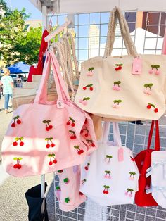 several purses are hanging up on a rack at an outdoor market, one is pink and the other is white with cherries