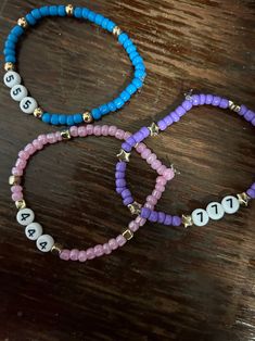 three bracelets with beads and letters on them sitting on top of a wooden table