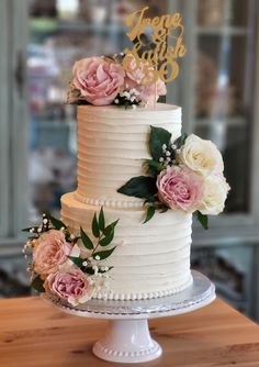 a white wedding cake with pink flowers and greenery on top, sitting on a wooden table