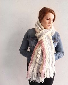 a woman wearing a white scarf with pink and grey fringes on the ends, standing against a wall