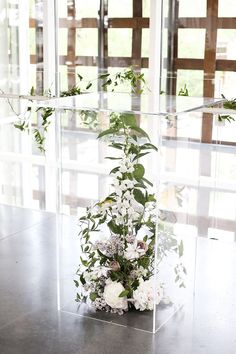 a vase filled with white flowers and greenery on top of a wooden table next to a glass wall