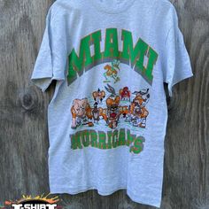 Vintage Ncaa Miami Hurricanes Looney Tunes T-Shirt, Miami Hurricanes Shirt, University Of Miami, Unisex T-Shirt Sweatshirt, Vintage Shirt Our Classic T-Shirt Serves As The Perfect Short-Sleeved Shirt For Your Unique, Funny, Or Personalized Designs. Brand: Gildan Heavy Weight Fabric Classic Unisex Makes This An Easy Fit Size Up If You Want Something Roomier Our Shirts Materials: 100% Cotton ** Note: - Double Check Your Address Before Ordering. - If You Want To Return The Goods, You Are The One To Crew Neck Tops With Front Print For Fan Gear, Fan Gear Graphic Tee With Crew Neck, Graphic Tee Tops With Fan Gear Graphic Print, Graphic Tee Tops With Graphic Print For Fans, Casual Green T-shirt For Fan Gear, Fan Gear Graphic Tee Shirt With Short Sleeves, Graphic Tee Shirt For Fan Gear With Short Sleeves, Gray Logo Print Top For Fan Merchandise, Short Sleeve Graphic Print Shirt For Fans