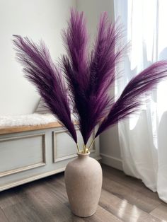 a white vase with purple feathers in it sitting on a wooden floor next to a window