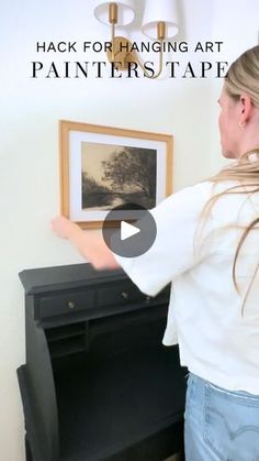 a woman standing in front of a painting on a black dresser with the words hack for hanging art painter's tape