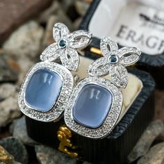 These classy dangle style pierced earrings are each accented with one (1), bezel set, cushion cabochon cut natural blue chalcedony, fifty-two (52), bead set, round brilliant cut diamonds and one (1), bezel set, round mixed cut natural blue sapphires. The earrings measure 28.0mm X 14.8mm and are finished with omega style backs. Elegant Blue Cabochon Earrings, Elegant Chalcedony Jewelry With Gemstone Accents, Elegant Chalcedony Drop Earrings, Elegant Chalcedony Jewelry For Anniversary, Formal Chalcedony Cabochon Jewelry, Formal Cabochon Chalcedony Jewelry, Elegant Blue Cushion Cut Earrings, Bead Set, Green Quartz