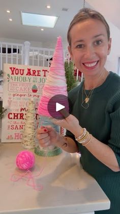 a woman is smiling and holding up a fake pink christmas tree in her hands, with balls of yarn on the table behind her