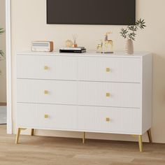 a white dresser with gold knobs in front of a pink wall and potted plant