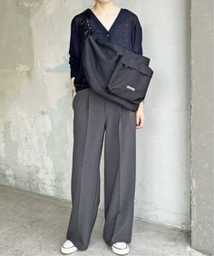 a woman standing in front of a concrete wall wearing grey pants and a black top