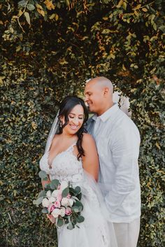 a bride and groom standing next to each other in front of a wall with greenery