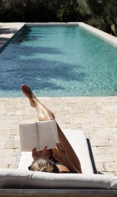 a woman laying on top of a white chair next to a pool reading a book