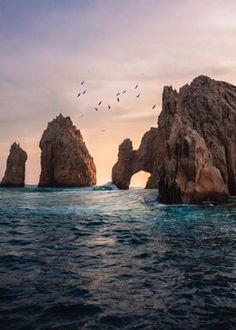 birds flying over the ocean near two large rocks