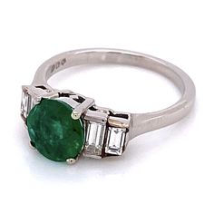 20th Century circa 1995 - Early Millennium era. Estate platinum ring featuring a center prong set natural emerald with each tiered step style shoulder channel set with two baguette cut diamonds.  The ring is finished with a bright polish and contain purity hallmarks from London, England. Gemstone: 1 round mixed cut emerald, 1.0 carat weight (calculated in mounting) medium tone strong green color (chip on girdle) Gemstones: 4 straight baguette step cut diamonds, 0.35 carats total weight (calculat Classic Formal Emerald Ring Channel Set, Classic Formal Emerald Ring With Channel Set, Formal Channel Set Emerald Ring, Classic Green Channel Set Emerald Ring, Formal Green Channel Set Emerald Ring, Channel Set Emerald Ring, Color Chip, Step Cut, Baguette Cut Diamond