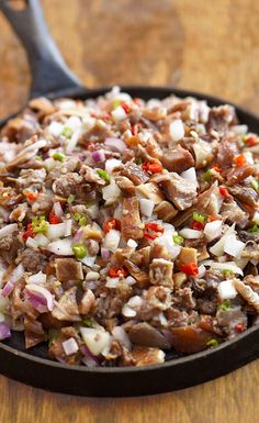 a skillet filled with meat and vegetables on top of a wooden table next to a spoon