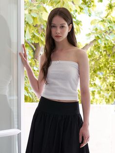 a woman standing in front of a window wearing a white top and black pleated skirt