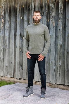 a man with a beard standing in front of a wooden fence wearing black jeans and a gray sweater