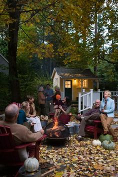 a group of people sitting around a fire pit in the middle of a yard with leaves on the ground