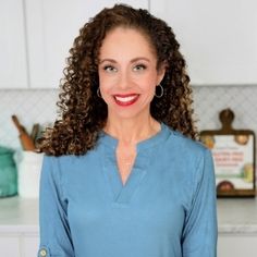 a woman with curly hair standing in front of a kitchen counter and smiling at the camera