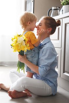 a woman sitting on the floor holding a bouquet of daffodils and kissing her baby