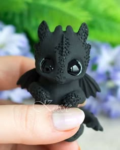 a tiny black dragon figurine sitting on someone's finger with flowers in the background