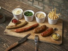 a wooden cutting board topped with sausages and fries next to two cups of dip