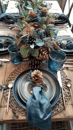 the table is set with pine cones, blue dishes and silver cutlery wares