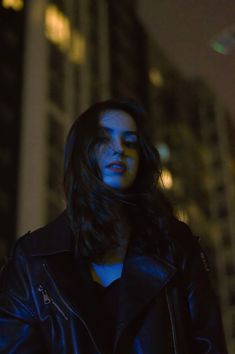 a woman with long hair standing in front of a tall building at night wearing a black leather jacket