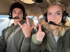 two people sitting in the cockpit of a plane with headphones on and one person pointing at the camera