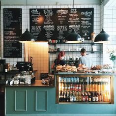 two people working behind the counter of a cafe with menus on the wall and lights hanging above it