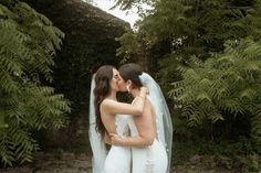 two brides are kissing in front of some trees and bushes on their wedding day