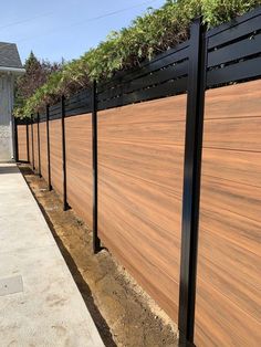 a wooden fence with black posts and plants growing on it's top, next to a sidewalk