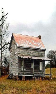 an old wooden house in the middle of a field with no grass and trees around it