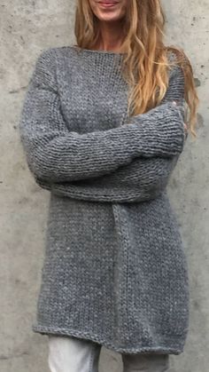 a woman standing in front of a concrete wall wearing a gray sweater and white jeans