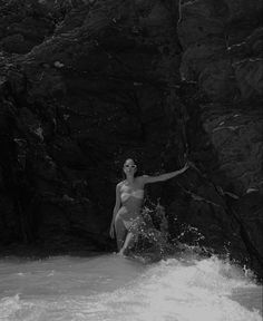 a woman standing in the water next to a rocky cliff and holding her arms out