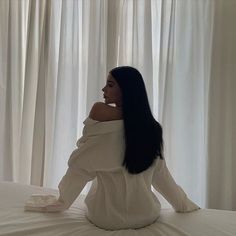 a woman sitting on top of a bed next to white curtains