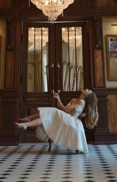 a woman in a white dress is sitting on a chair and looking up at the chandelier