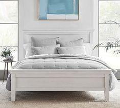 a white bed sitting in a bedroom next to two windows and a painting on the wall