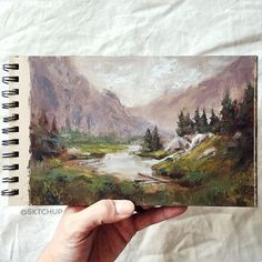 a person holding up a book with an image of mountains in the background and a river running through it