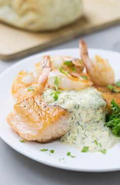 a white plate topped with fish and shrimp covered in cream sauce next to bread on a cutting board